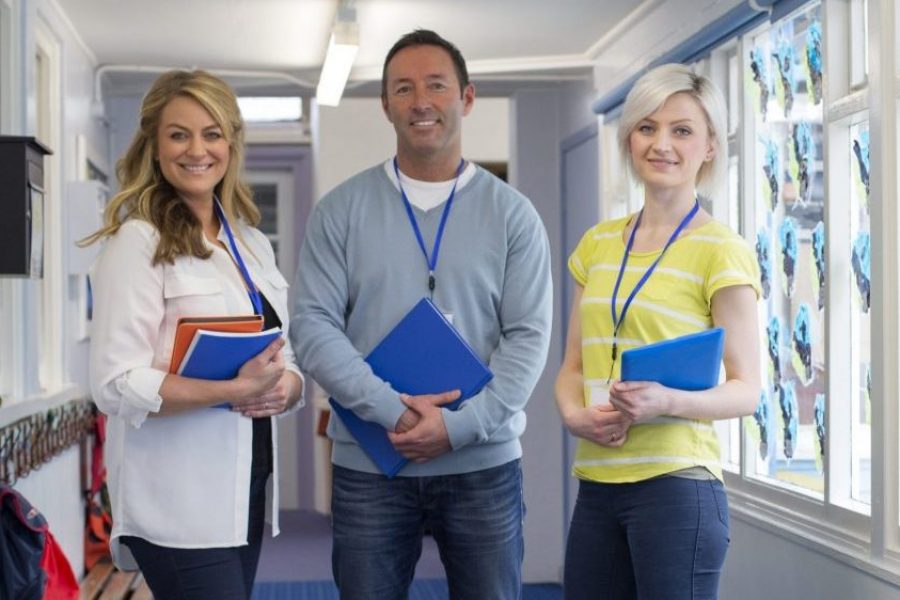 Three teachers standing in the corridor of a school building. They are holding school work and smiling at the camera.