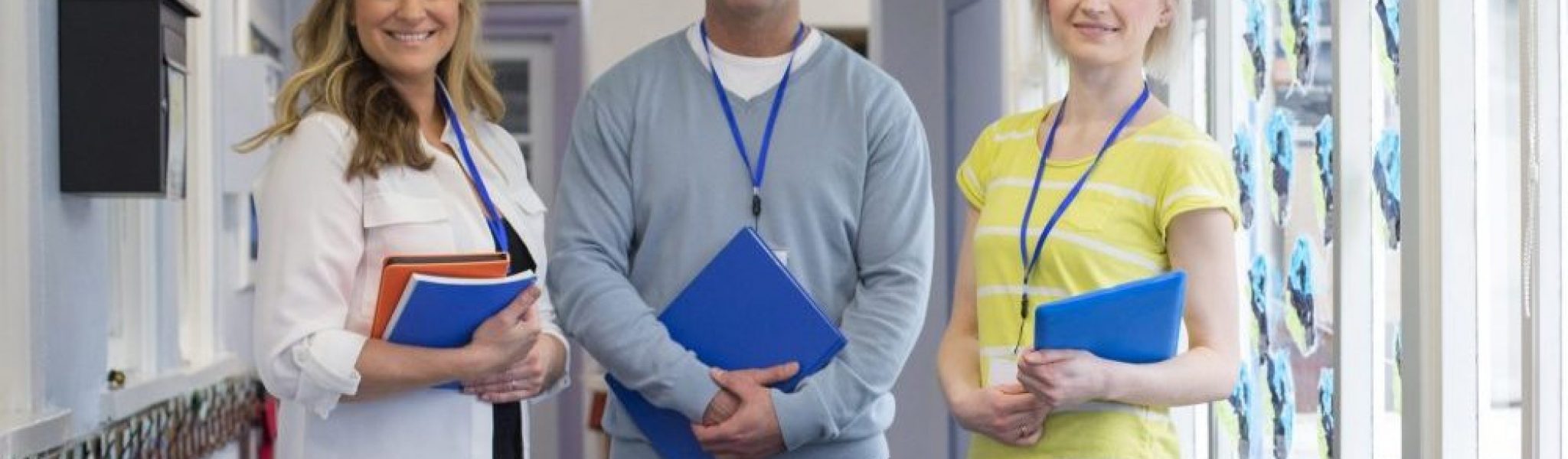 Three teachers standing in the corridor of a school building. They are holding school work and smiling at the camera.