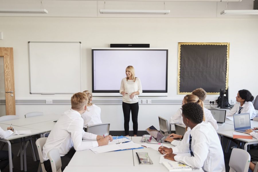 Female High School Teacher Standing Next To Interactive Whiteboard And Teaching Lesson To Pupils Wearing Uniform