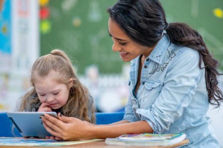 A young teacher shows her student something on a tablet at her classroom desk. Her student is elementary aged and has Down syndrome. She shows interest in the tablet.