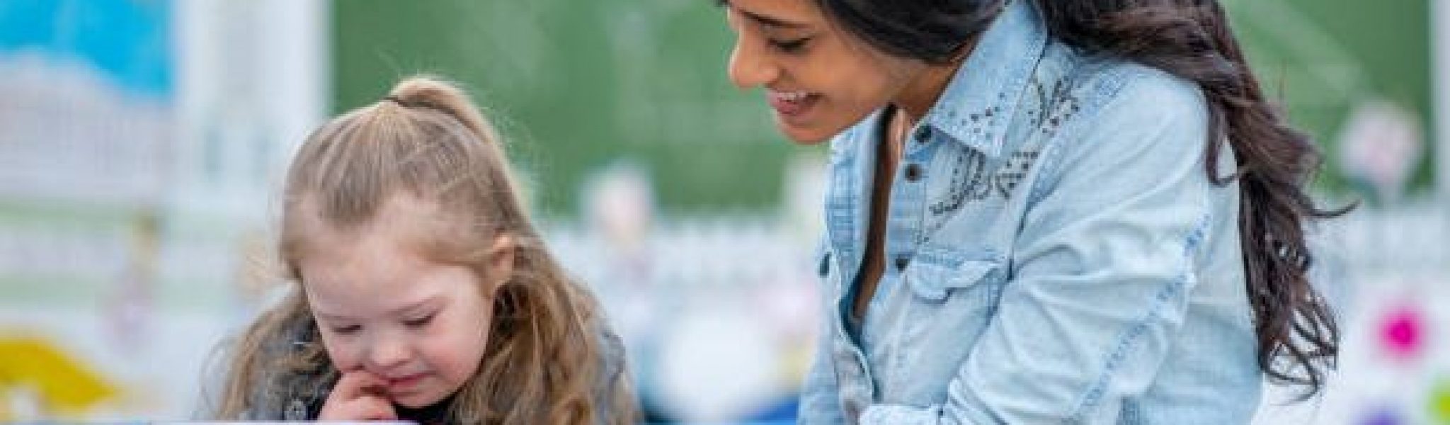 A young teacher shows her student something on a tablet at her classroom desk. Her student is elementary aged and has Down syndrome. She shows interest in the tablet.