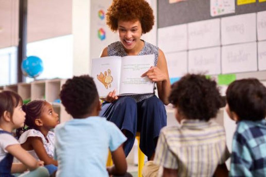 Female Teacher Reads To Multi-Cultural Elementary School Pupils Sitting On Floor In Class At School